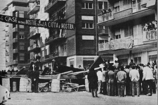 A black and white photograph of an Italian working class estate in the 1970s. A banner hangs between building with the words 'Le cases sono nostre, Tutta la citta e nostra'